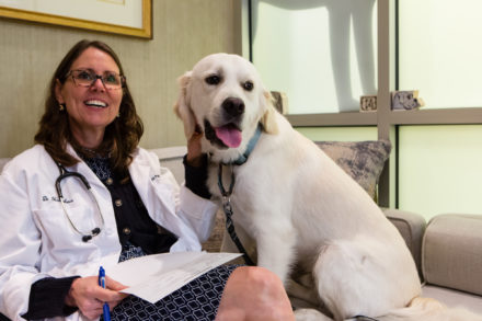 A smiling veterinarian sits on a couch next to a happy dog